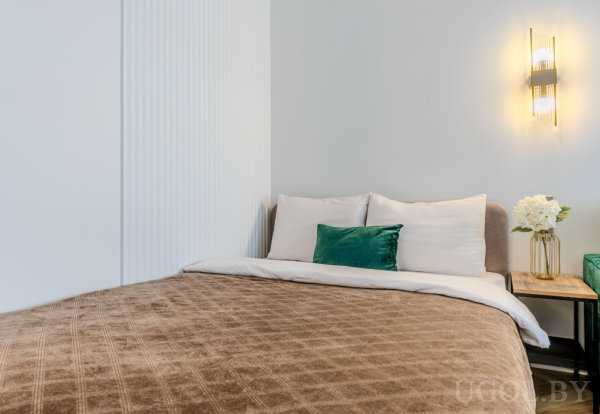 Кровать с коричневым покрывалом, белыми подушками и зелёным акцентом, тумба с вазой и цветами, настенный светильник и светлая декоративная панель.
