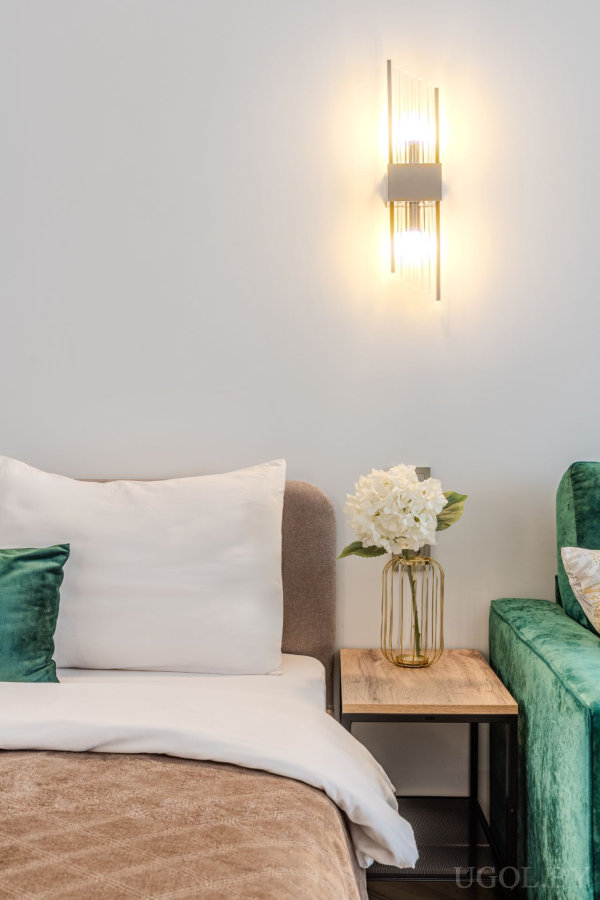 Кровать с бежевым изголовьем, белыми и зелёными подушками, тумба с вазой и цветами, зелёное кресло и настенный светильник с вертикальными стеклянными элементами.