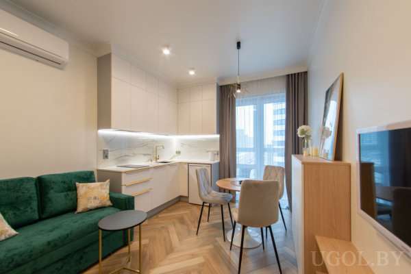 Кухня с белыми фасадами и мраморным фартуком, круглый деревянный стол с мягкими стульями, зелёный диван и тумба с декором у стены.