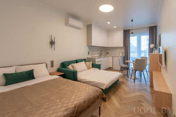 Комната с кроватью и зелёным диваном, кухней с белыми фасадами, круглым столом и большими окнами с тюлем на фоне паркетной ёлочки.