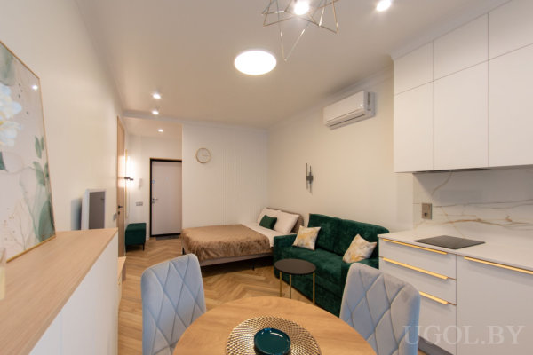 Кухня с белыми шкафами и мраморным фартуком, круглый стол с мягкими стульями, зелёный диван и кровать в глубине комнаты.