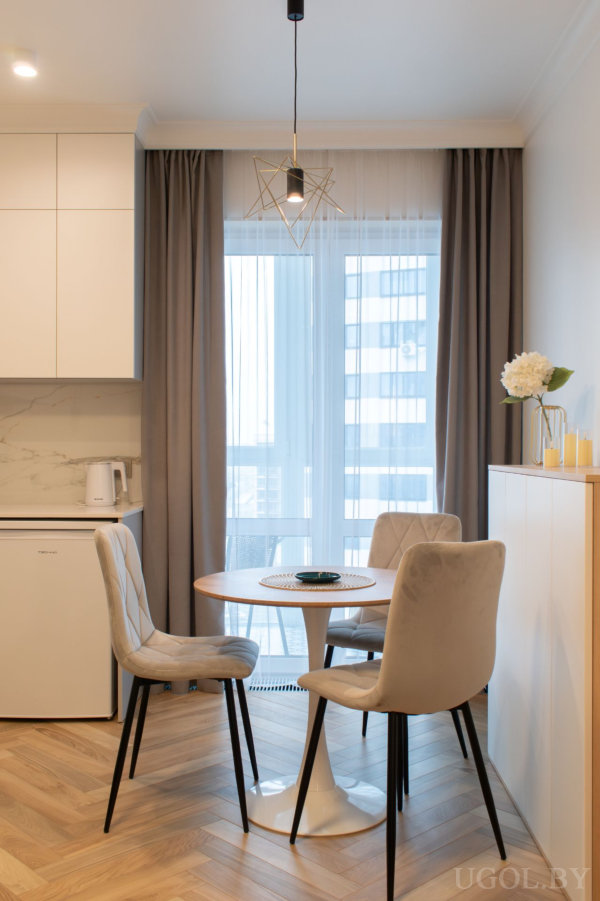 Круглый деревянный стол со светлыми стульями, окно с тюлем и серыми шторами, рядом белая тумба с декором и часть кухни с белыми шкафами.