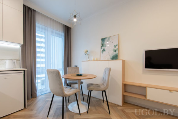 Круглый деревянный стол с серыми стульями, белый комод с декором, телевизор на стене и светлый паркет ёлочкой у окна с тюлем.