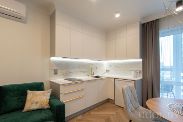 Белая кухня с мраморным фартуком и золотой фурнитурой, рядом зелёный диван, круглый стол и светлый паркет в студийном пространстве.