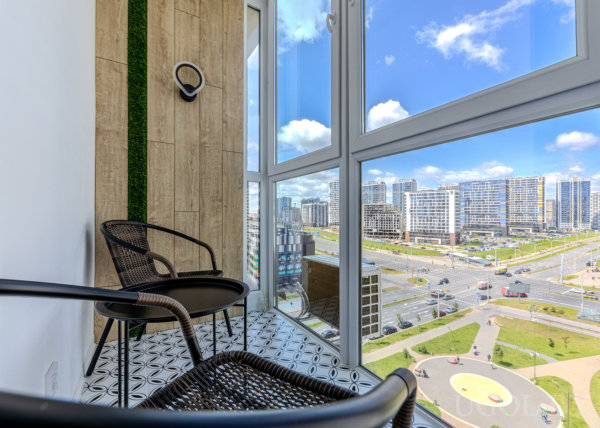 Застеклённая лоджия с панорамным видом на город, двумя плетёными креслами, круглым столиком и узорной плиткой на полу.