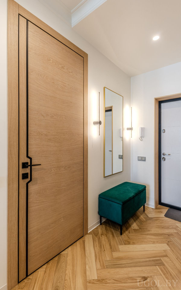 Стильная прихожая с деревянной дверью с чёрным декором, зеркалом в золотой раме, настенными светильниками и бирюзовой банкеткой.