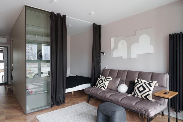 Современная студия с серым диваном, журнальным столиком, пуфом и зоной сна за шторами с кроватью и шкафом.