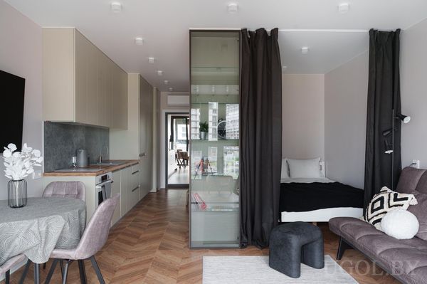 Компактная студия с круглым столом и розовыми креслами, кухней с бежевыми шкафами и зоной сна за стеклянной перегородкой.