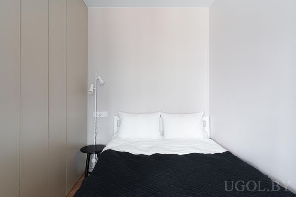 Минималистичная спальня с кроватью в чёрном покрывале, белыми подушками, прикроватным столиком и высоким бежевым шкафом.