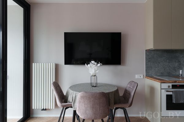 Небольшая обеденная зона с круглым столом, розовыми креслами, вазой с цветами и телевизором на стене.