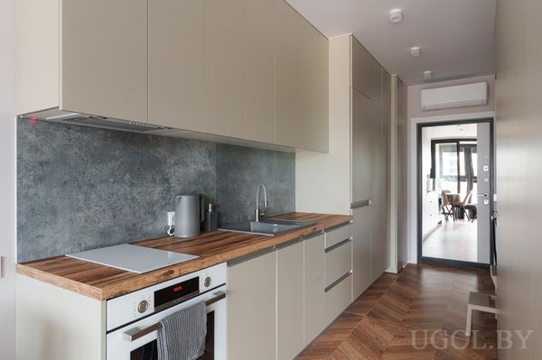 Современная кухня с бежевыми шкафами, деревянной столешницей, плитой, мойкой и серым фартуком.