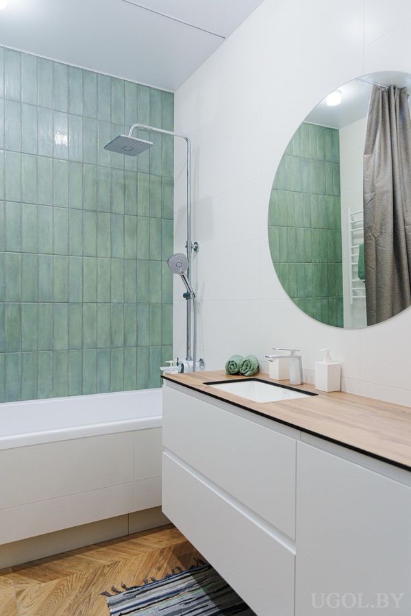 Современная ванная с белой тумбой, круглым зеркалом, зелёной плиткой и ванной с душем.