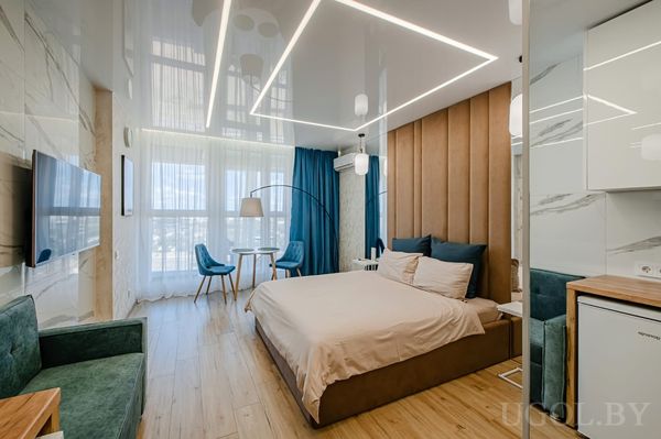 Современная студия с большой кроватью, мягким изголовьем, зелёным креслом, телевизором на мраморной стене и кухонной зоной с белыми шкафами.