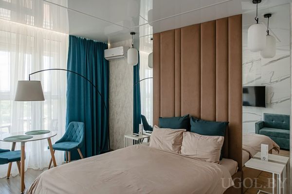 Стильная спальня с высоким мягким изголовьем, бордовым покрывалом, прикроватными тумбами и зоной отдыха с креслами и столиком у окна.