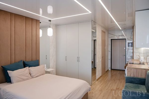 Компактная студия с кроватью у мягкого изголовья, белыми шкафами, кухней с деревянной столешницей и зеркалом у входа.