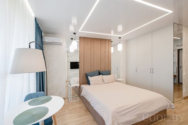 Современная спальня с кроватью, подвесными светильниками, круглым столиком с двумя креслами и телевизором на стене.