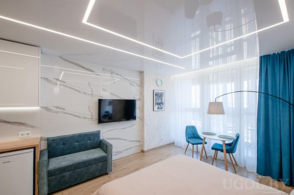 Студия с бирюзовым диваном, телевизором на стене, круглым столиком с креслами у окна и глянцевым потолком с подсветкой.