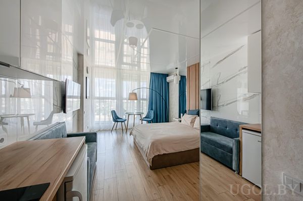 Современная студия с кроватью, мягким синим изголовьем, диваном, кухней с белыми шкафами и обеденной зоной у окна.