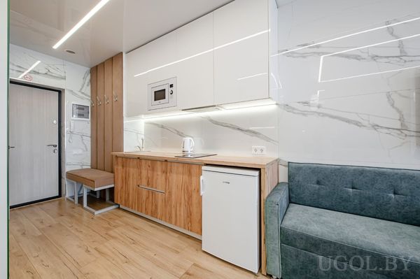 Компактная кухня с белыми шкафами, микроволновкой, чайником, диваном и прихожей с вешалкой и скамьёй.