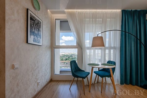 Современная обеденная зона с круглым столом, двумя бирюзовыми креслами, торшером и панорамным видом на город.