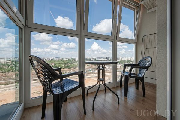 Застеклённый балкон с двумя чёрными креслами, круглым столиком и видом на железную дорогу и город.