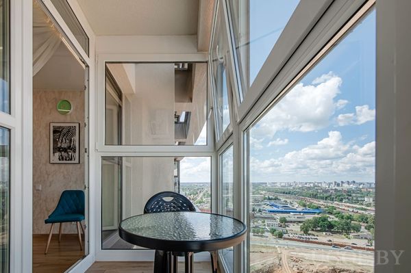 Современный балкон с круглым стеклянным столиком, креслом и видом на город, видна часть комнаты с креслом и светильником.