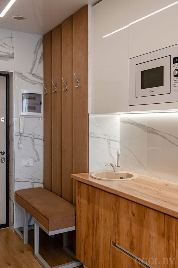 Компактная кухня с белыми шкафами, мойкой, микроволновкой и прихожей с мягкой скамьёй и вешалкой.