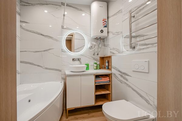 Современная ванная с угловой ванной, белой тумбой с круглым зеркалом, водонагревателем и полотенцесушителем.