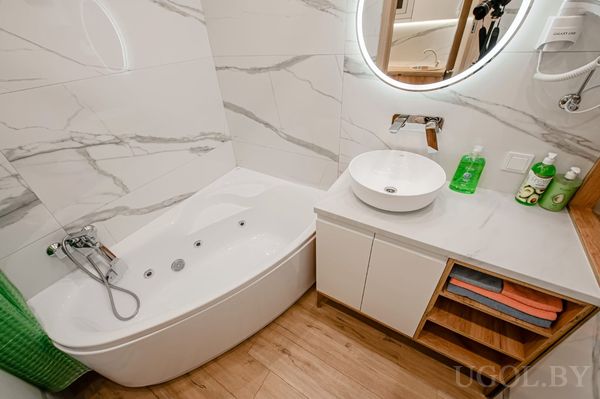 Компактная ванная с угловой ванной, круглым зеркалом с подсветкой, белой тумбой и открытыми полками с полотенцами.