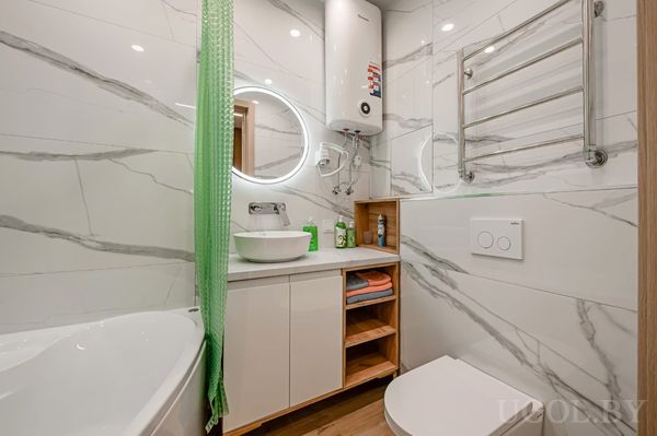 Современная ванная с круглым зеркалом с подсветкой, белой тумбой, зелёной шторой и водонагревателем.