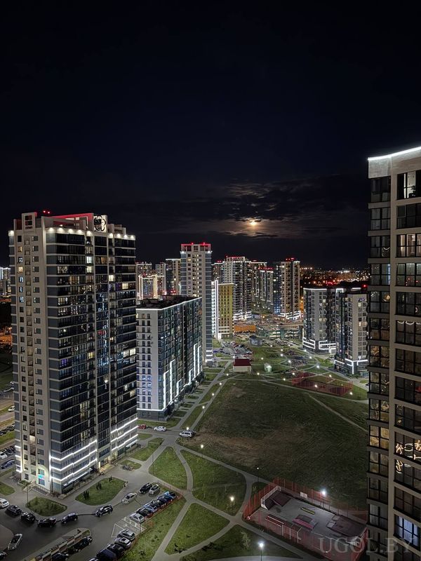 Ночной городской пейзаж с многоэтажными домами, подсветкой фасадов и луной в облаках.