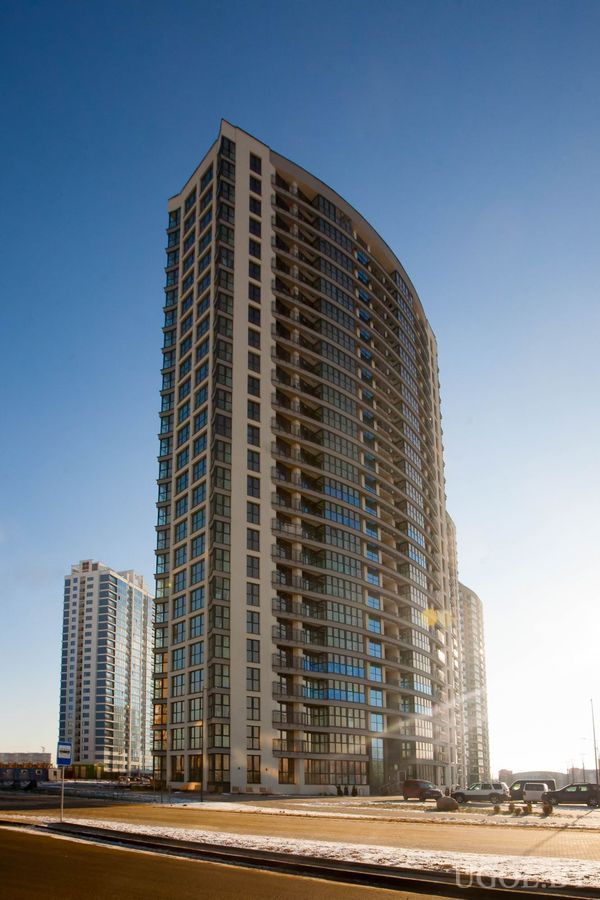 Современный жилой дом с изогнутым фасадом, балконами и стеклянными окнами, на переднем плане дорога и снег.