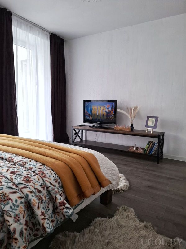 Современная спальня с цветочным покрывалом, ковром, телевизором на консоли с декором и большим окном.