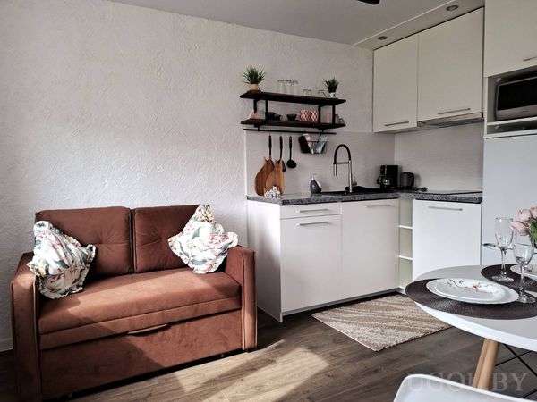 Компактная кухня с белыми шкафами, чёрной столешницей, диваном с цветочными подушками и открытыми полками.