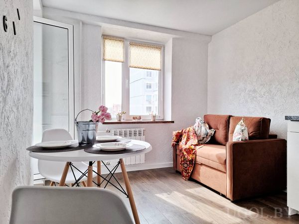 Уютный уголок с круглым столом, орхидеей, креслом с пледом и большим окном с жалюзи.