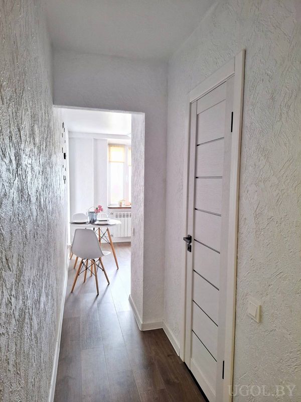 Светлый коридор с белой дверью, тёмным полом и видом на столовую с сервированным столом у окна.