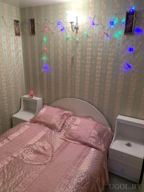 Комната с кроватью, белыми тумбами, лампой, гирляндой, полосатыми обоями и декоративными картинками.