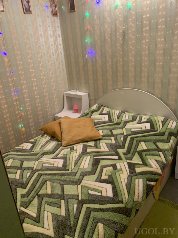 Интерьер с кроватью, подушками, тумбой, лампой, гирляндой и картинками на фоне полосатых обоев.