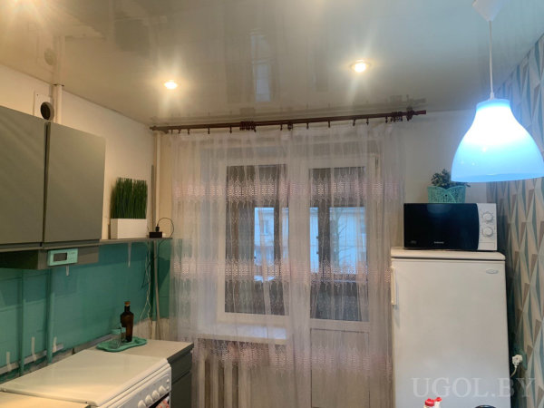 Рабочая зона с плитой, холодильником, микроволновкой, растением, подсветкой и белыми шторами.