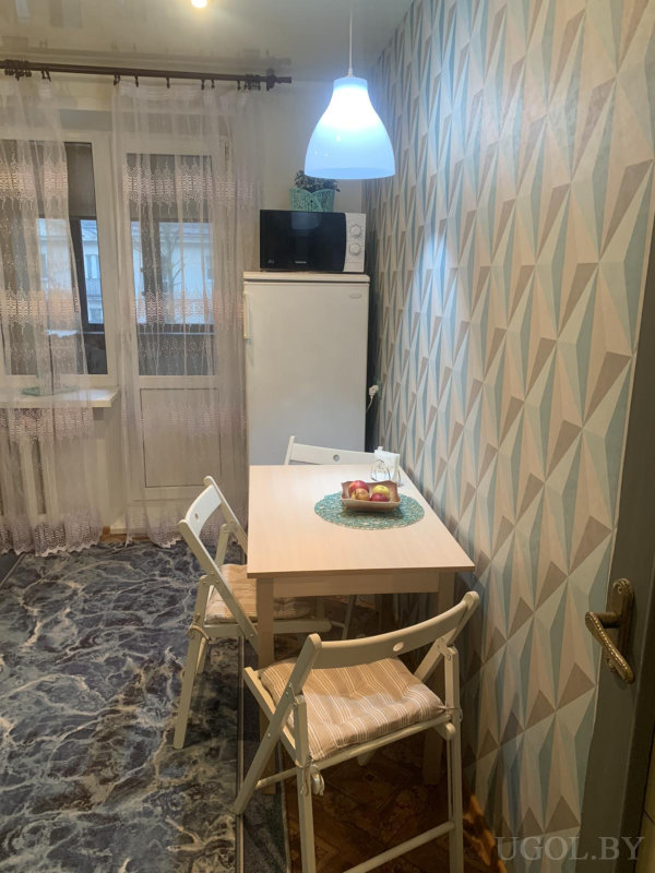 Комната с столом, складными стульями, ковриком, холодильником, микроволновкой и балконной дверью.