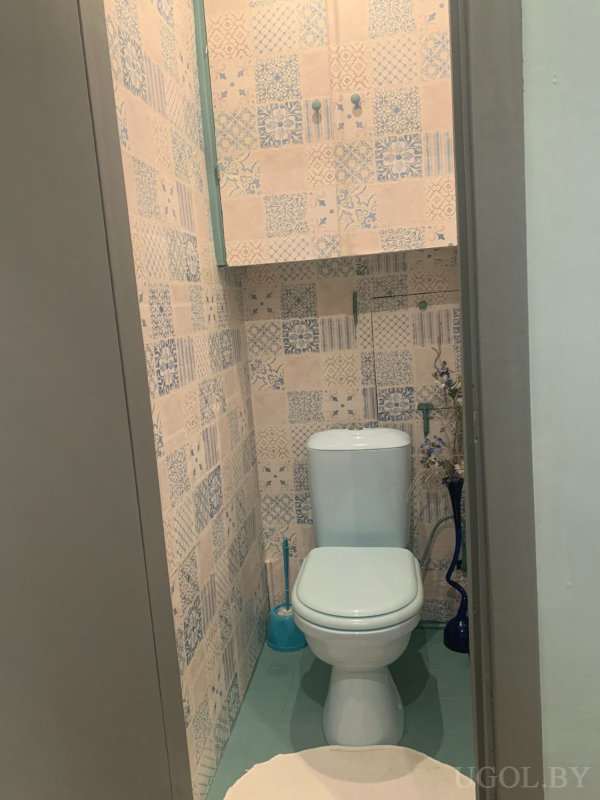 Комната с унитазом, шкафчиком, вазой с цветами, ёршиком и плиткой с орнаментом.