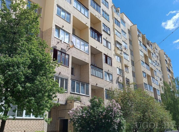 Уличная сцена с многоэтажным домом, балконами, спутниковыми антеннами и зеленью на переднем плане.