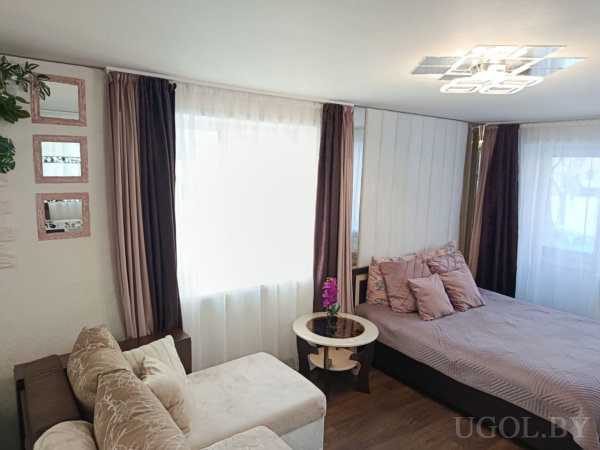 Кровать с сиреневыми подушками, круглый столик с фиолетовой орхидеей, бежевый диван и окно с тюлем и тёмными шторами.