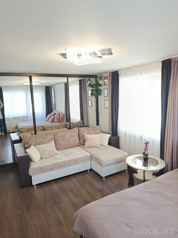 Бежевый диван, стеклянный столик с цветами, зеркальные панели, окно с тюлем и фрагмент кровати на переднем плане.