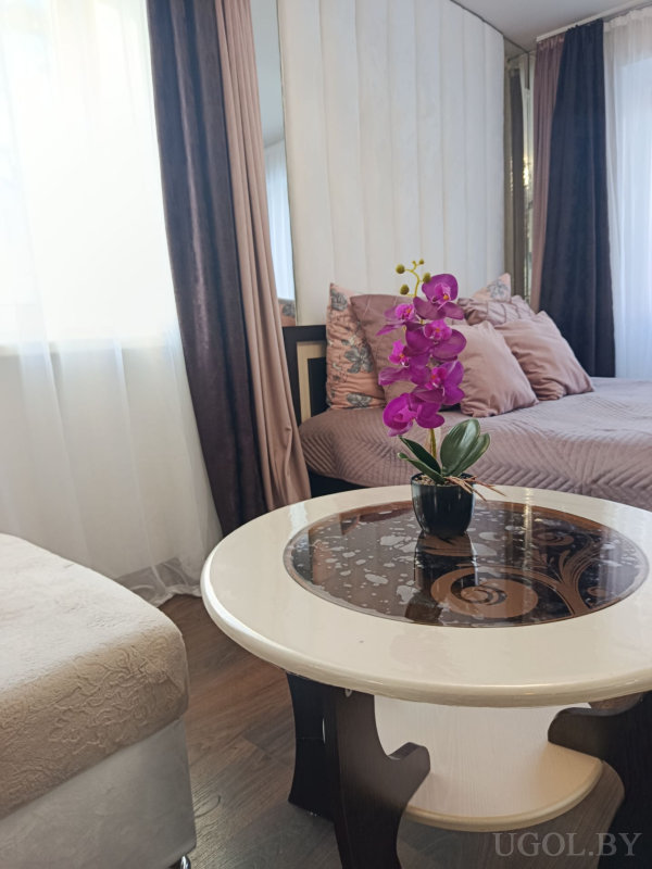 Кровать с сиреневым покрывалом, круглый стол с узором и орхидеей, вертикальные панели на стене и мягкий пуф.