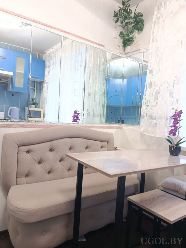 Бежевый диван, деревянный стол с чёрными ножками, зеркало, голубые шкафы и подвесное растение в углу.