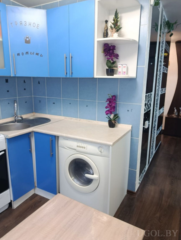 Голубые шкафы, встроенная стиральная машина, плитка с узором, декоративные элементы и орхидеи на полках и столешнице.