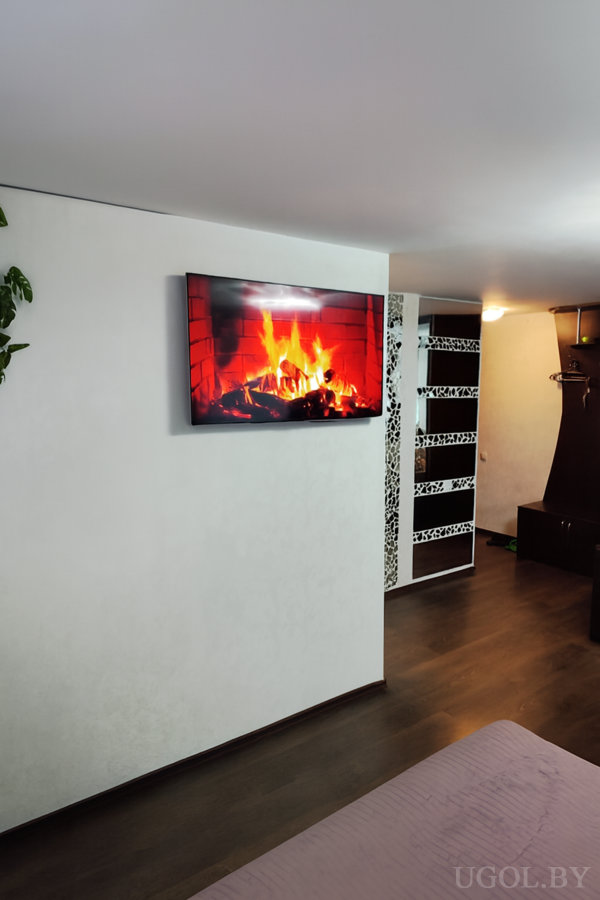 Телевизор с изображением огня, белая стена, чёрно-белая перегородка, фиолетовое покрывало и вешалка на фоне.