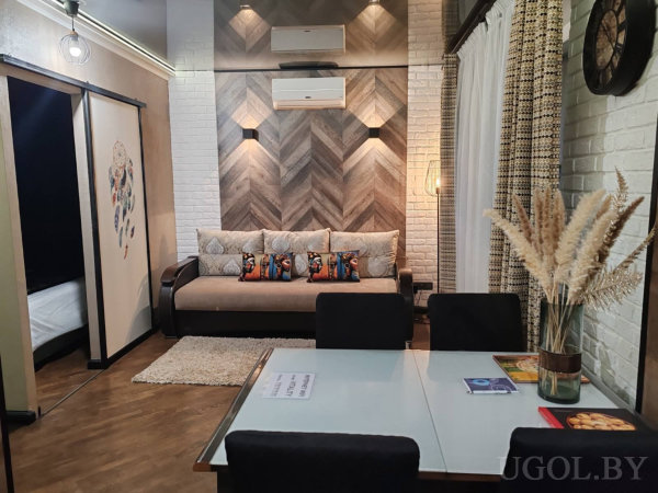 Диван с декоративными подушками на фоне деревянной панели, рядом обеденный стол со стеклянной столешницей и чёрными стульями, ваза с сухоцветами и вид на спальню через раздвижную дверь.