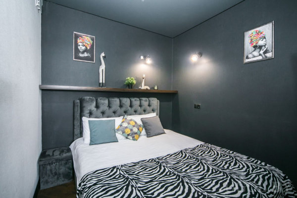 Кровать с мягким серым изголовьем, белым бельём и зебровым покрывалом, над ней полка с фигуркой жирафа и растением, тёмные стены с яркими портретами.